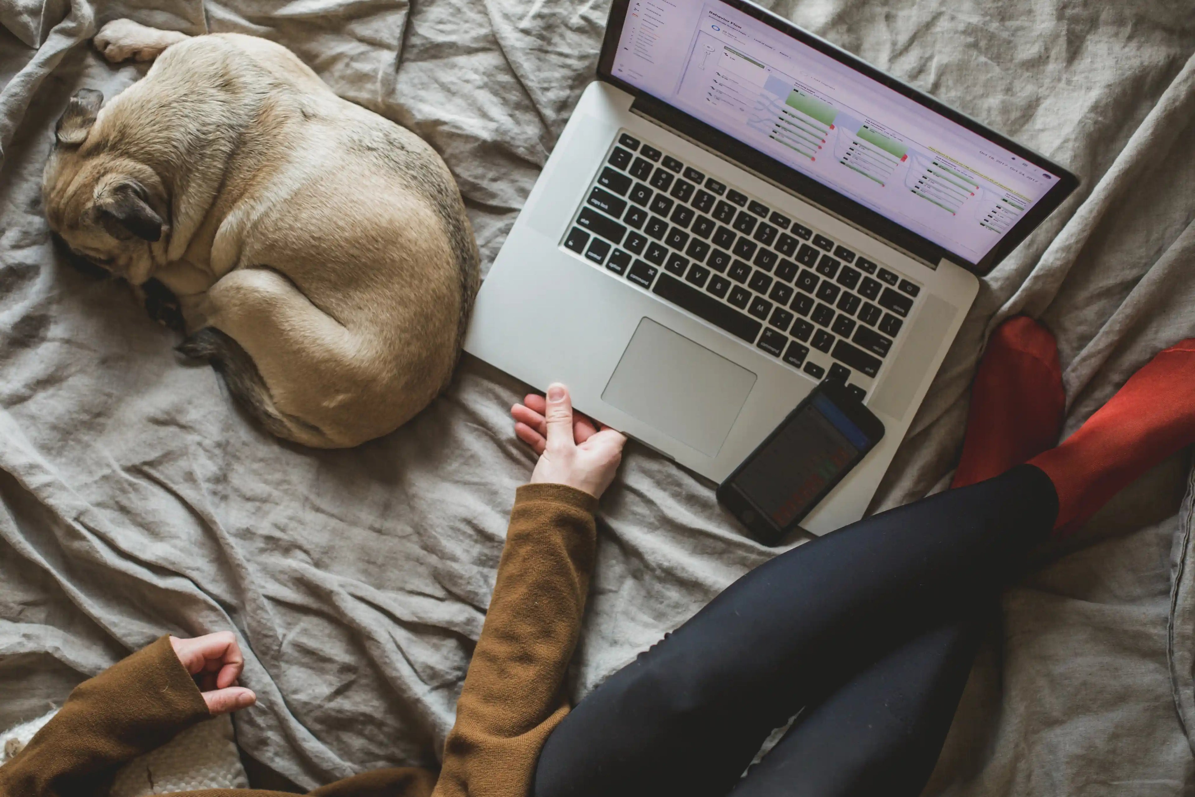 En dame som jobber hjemmefra med en laptop, mobiltelefon og en hund som ligger ved siden av.