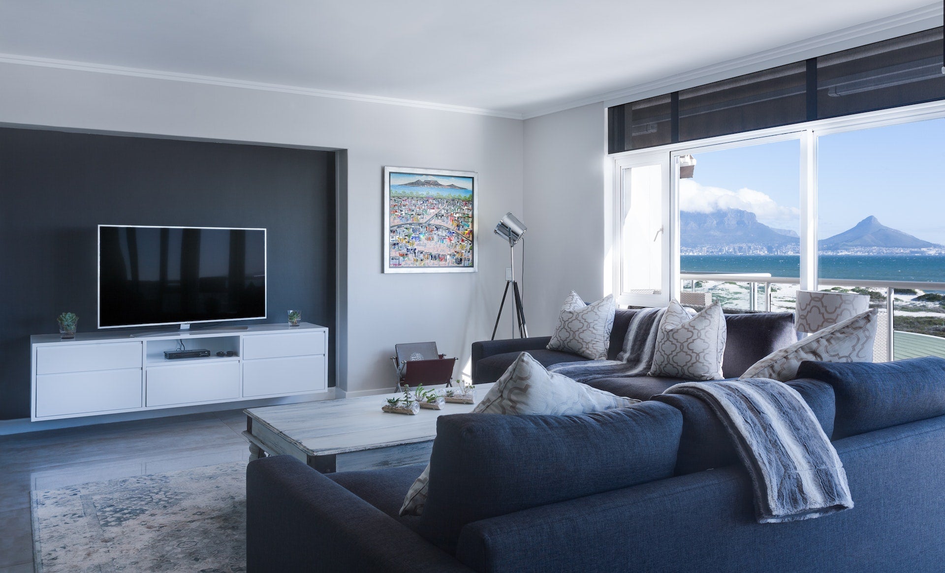 En moderne stue med en stor sofa, TV, pynteputer, pledd og en vakker utsikt mot havet.
